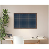 Navy blue framed fabric pinboard with thin white grid lines mounted on beige wall above a light wood desk, accompanied by a leafy green plant and a white chair.