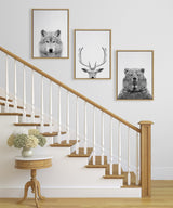 Three black and white animal portraits—a wolf, a deer with antlers, and a bear—framed and hung in a descending diagonal line alongside a wooden staircase with white balusters.