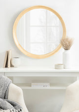 Round wall mirror with a smooth light wood frame reflecting a window, mounted above a white shelf holding books, a white vase with dried grasses, and a small round container.