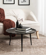 Round black coffee table with a scalloped edge and tapered legs, topped with two black vases holding dried branches and two stacked books, placed on a patterned beige rug.