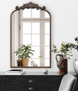 Arched mirror with dark carved wood frame featuring floral motifs, reflecting a window and two potted green plants placed on a black cabinet with books and decor.