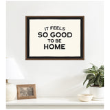 Framed black and white wall art with bold typography reading 'It feels so good to be home,' displayed above a white shelf with a lamp, photo frame, plant, and bowl.