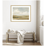 Framed landscape print of rolling beige hills under a soft cloudy sky, hanging above a wooden bench with an open book and draped white cloth in a serene, minimal room.