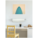 Minimalist workspace with a wooden chair and white desk, featuring a framed teal surfboard tip artwork on a light gray wall and a roll of white paper beneath it.