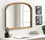Arched wall mirror with a warm wood frame above a white desk featuring potted plants, books, and black-and-white photos on an adjacent wall.