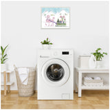 Framed canvas artwork showing a pink beaded poodle pulling a sled with presents against a snowy mountain backdrop, hanging above a white front-load washer in a minimalist laundry room.