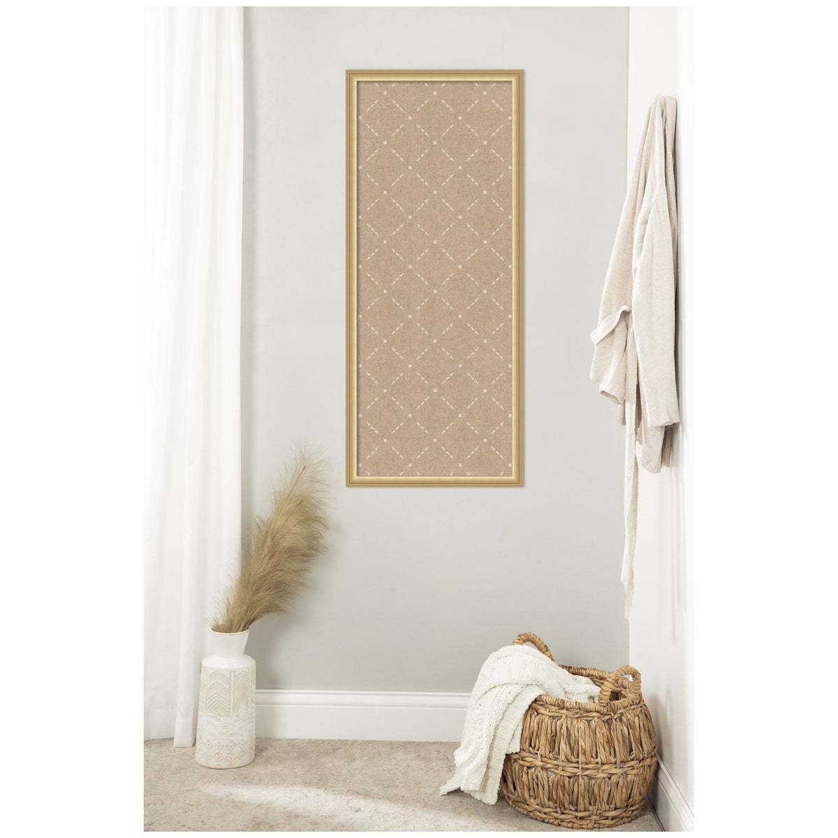 Vertical rectangular beige pinboard with a white lattice pattern framed in light wood, hung on a light gray wall above a wicker basket with a white cloth and a white vase holding dried pampas grass