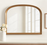 Arched wall mirror with a warm brown wood frame, reflecting a window and a bright, softly lit room corner, resting above a white vase with cream-colored flowers.