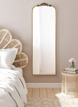 Tall rectangular arched mirror with ornate gold detailing on top, mounted on a beige wall beside a rattan headboard and a woven side table with flowers and books.