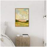 Framed canvas painting of a soft landscape with a cloudy sky in pastel blues, yellows, and greens hanging on a white wall over a rustic wooden side table beside a white bed.