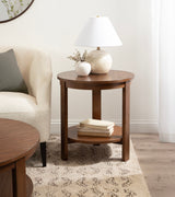 Round wood side table with a lower shelf holding books, topped with a white ceramic lamp and a small vase against a light neutral wall and sheer curtain.