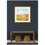 Square framed painting of a colorful meadow with yellow fields, pink and red flowers, and a light blue sky above, hanging on a dark gray wall over a wooden bench.