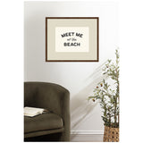 Square framed wall art with 'MEET ME at the BEACH' black text on white background, hanging above an olive green curved armchair and a potted olive tree in a woven basket.