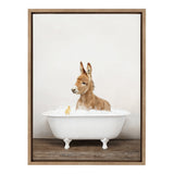 Young brown donkey sitting in a white clawfoot bathtub filled with bubbles, accompanied by a small yellow rubber duck, set against a plain light gray wall and wooden floor.