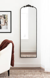 Tall, narrow wall mirror with arched top and ornate black decorative frame, mounted in a white room above a multi-toned woven rug and beside a cream sofa with a brown knitted throw.