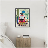Framed colorful illustration of a black cat sitting on a pink chair reading a newspaper, surrounded by potted plants and a blue lamp, hung on a white wall above a wooden bedside table with a cup and books.