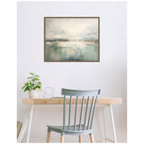 Framed abstract canvas with gray and green horizon tones hanging above a minimalist wooden desk and gray spindle-back chair, with a small plant and decorative wire sphere on the desk.