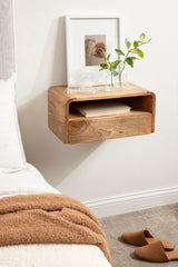 Wall-mounted floating side table in smooth natural wood with rounded edges, featuring an open shelf holding a book and a top surface with two glass vases and a framed photo against a white wall.