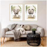 Two framed prints above a sofa depict an elephant in a bathtub with green palm leaves, shown front and back view in neutral tones with light wood frames.