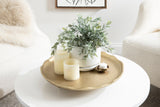 Round gold decorative tray with a scalloped edge holding three white pillar candles, a small candle in a gold holder, and a white pot with green foliage on a white round table