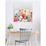 Square canvas painting of vibrant mixed flowers in white, pink, yellow, and red hues with green leaves on a soft gray background, hanging above a wood desk.