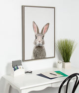 Framed canvas print of a young gray rabbit with upright ears hanging on a white wall above a white desk featuring a green plant, owl figurine, and office supplies.