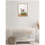 Framed canvas of a baby elephant sitting in a green clawfoot bathtub with white walls reflected behind, hanging above a white bench with a fur throw and a bag on the floor.