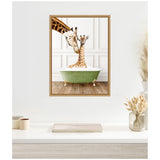 Framed canvas showing a mother giraffe nuzzling a baby giraffe sitting in a green clawfoot bathtub filled with bubbles against a white paneled wall.