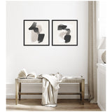 Two framed abstract canvas artworks with overlapping black and beige irregular shapes, hung side by side above a wooden bench with an open book and draped fabric in a light, airy room.