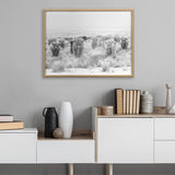 Framed black and white photo of a herd of Highland cows standing in a grassy field, hanging on a gray wall above a modern white and wood sideboard with vases and books.