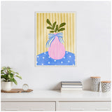 Framed canvas painting of a pink vase with green leaves and a blue ribbon on a blue polka-dot surface, set against a vertical yellow striped background.