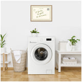 White front-loading washing machine centered between woven laundry basket with a white towel and a white table holding detergent and folded towels, with plants and a framed vintage postcard reading 'It feels so good to be home' on