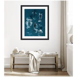 Framed aerial art of a school of humpback whales swimming in deep blue ocean water, displayed above a simple wooden bench with a draped white cloth.