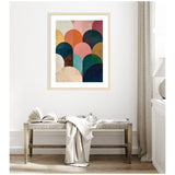 Framed minimalist geometric abstract art with overlapping jewel tone semicircles in warm reds, blues, and golds, hung above a wooden bench with an open book and draped fabric.