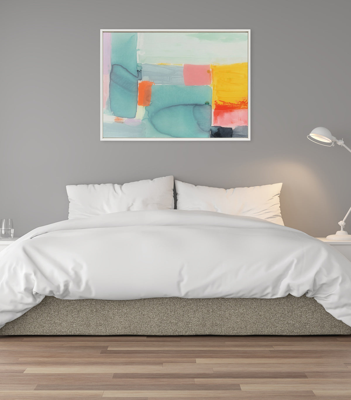 Abstract painting with teal, coral, yellow, and gray color blocks in a white frame hanging above a white bed against a gray wall, with a white desk lamp on the right.