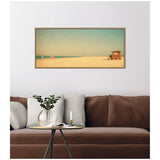 Framed panoramic photo of a sandy beach with a small wooden lifeguard hut near the right edge, under a clear blue sky with a gradient to a warm horizon.