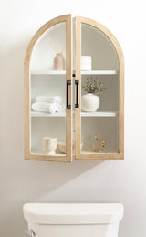 Wall-mounted wooden arch cabinet with glass doors, black handles, showcasing white shelves holding rolled towels, vases, and decorative items against a light wall.