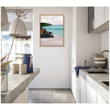Framed painting of a seascape with turquoise water, black rocks, and green hills, hanging in a bright white kitchen with gray countertops and ceramic mugs.