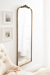 Tall, arched wall mirror with an ornate bronze frame and decorative scrollwork at the top, positioned beside beige tufted chair and framed art on a white wall.