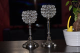 Two silver candle holders with spherical tops made of clear crystal beads arranged in a cluster, displayed on a dark surface with a white pot plant beside them.