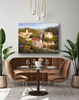 Painting of a sunlit European village with pastel-colored buildings and a church tower, displayed above a round glass table with brown velvet chairs and green plants.