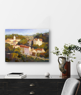 Painting of a sunlit hillside town with clustered buildings among green trees under a blue sky, displayed on a white wall above a black desk with books and plants.
