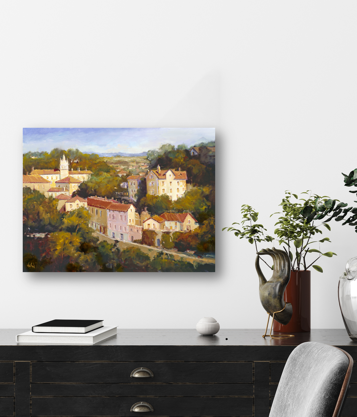 Painting of a sunlit hillside town with clustered buildings among green trees under a blue sky, displayed on a white wall above a black desk with books and plants.