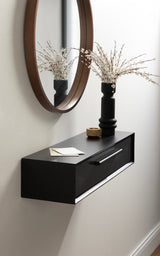 Black floating console table with a drawer, mounted on a white wall under a round brown-framed mirror, holding a black vase with white dried flowers and a small wooden bowl.