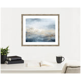 Abstract framed artwork featuring blue, gray, and beige tones blending horizontally, displayed above a white desk with books, plant, open notebook, and black mug.