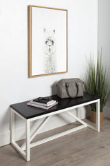 Minimalist room corner with a black-topped bench featuring white metal legs, holding a gray bag and books, beneath a framed alpaca print on a white wall, next to a tall green plant.