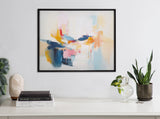 Abstract coastal-themed painting with blue, pink, orange, and yellow brushstrokes hanging above a white surface decorated with plants, books, and a salt lamp.