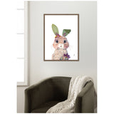 Watercolor-style bunny portrait with large eyes and floral accents including green leaves as ears and purple flowers below the chin, framed in wood on a white wall above an olive-green armchair with a knitted cream throw.