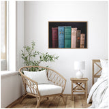 Framed canvas featuring a row of vintage classic books in muted red, blue, green, and brown tones hanging on a white wall above a beige wicker chair and wooden nightstand in a minimalist room.