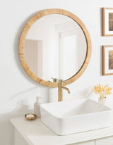 Round wall mirror with textured natural wood frame above white countertop holding a square white vessel sink and brass faucet, flanked by soap dispenser and dried floral arrangement.